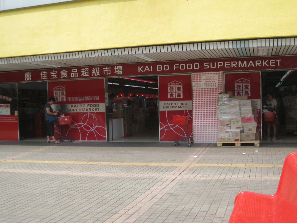 The image depicts the entrance of Kai Bo Food Supermarket in Hong Kong, with a few shoppers and stacks of boxes near the doorway.